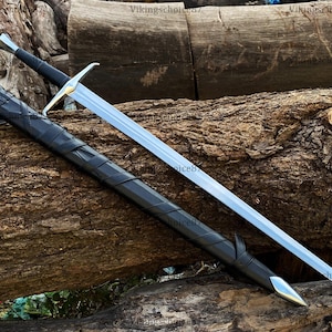 May include: A long, silver sword with a gold crossguard and black handle rests beside its black leather sheath. The sword and sheath are displayed on a textured, brown log, showcasing a medieval or fantasy theme.