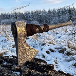 May include: A silver axe with intricate carvings and a brown leather-wrapped handle. The axe head features detailed patterns and a circular design. The axe is resting on a piece of wood in a snowy outdoor setting.