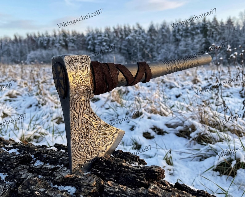 May include: A silver axe with intricate carvings and a brown leather-wrapped handle. The axe head features detailed patterns and a circular design. The axe is resting on a piece of wood in a snowy outdoor setting.