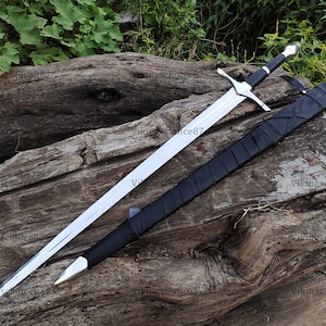 May include: A long, silver sword with a cross-shaped hilt and a black handle, resting on a weathered wooden surface. A black sheath accompanies the sword. The blade is sharp and the design is detailed.