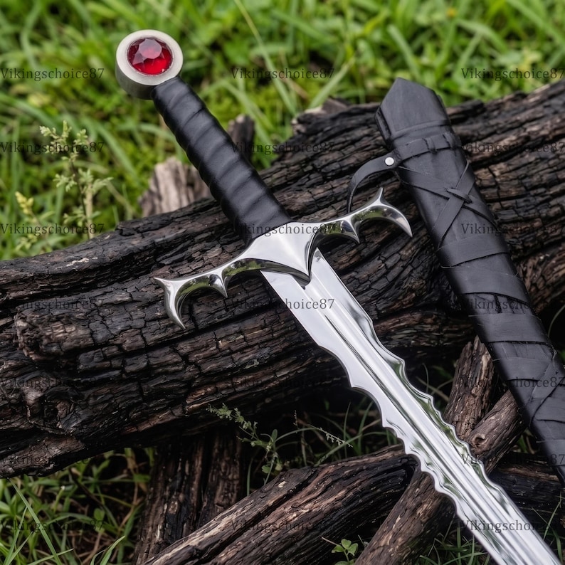 May include: A silver-coloured metal sword with a wavy blade, a black leather-wrapped handle, and a pommel featuring a circular red gemstone. The sword includes a matching black leather scabbard with crisscross lacing details along the side.
