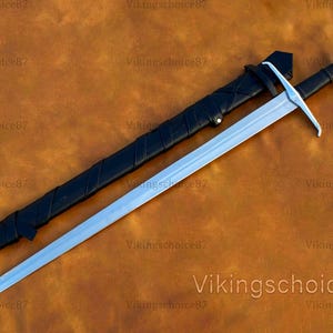 May include: A long, silver sword with a black handle and a black sheath. The sword has a cross-shaped hilt and a pointed tip. The sheath is lying next to the sword on a brown surface. The text "Vikingschoice87" is visible.