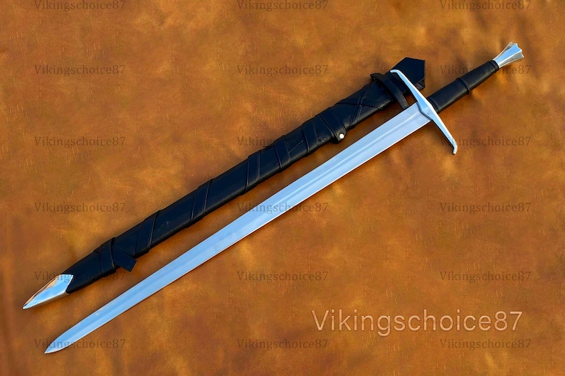 May include: A long, silver sword with a black handle and a black sheath. The sword has a cross-shaped hilt and a pointed tip. The sheath is lying next to the sword on a brown surface. The text "Vikingschoice87" is visible.
