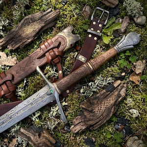 May include: A detailed image of a Viking sword with a brown leather sheath and belt. The sword features an ornate handle and blade with a green gem. The sheath and handle are intricately carved with patterns. The background is moss and wood.