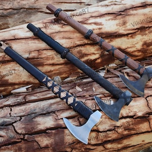 May include: Three Viking axes with black leather wrapped handles and different colored wooden shafts. The axes have different shaped heads and are displayed on a wooden background.