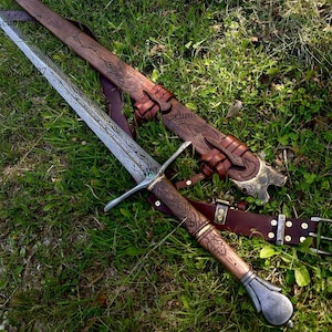 May include: A detailed image of a longsword with a patterned blade and wooden handle, resting on grass. The sword is accompanied by a brown leather sheath and belt, featuring intricate carvings and metal accents. The overall aesthetic is reminiscent of historical weaponry.