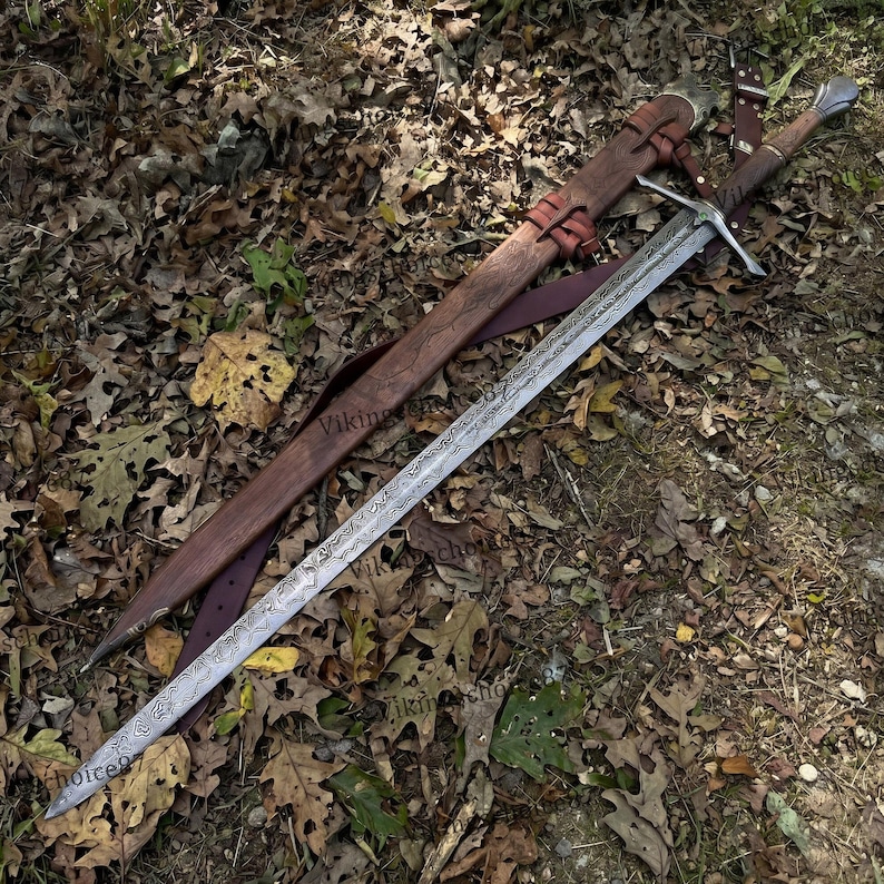 May include: A long, ornate sword with a patterned blade and a wooden handle, resting on a bed of fallen leaves. The sword is accompanied by a brown leather sheath with red leather straps. The overall design suggests a historical or fantasy theme.