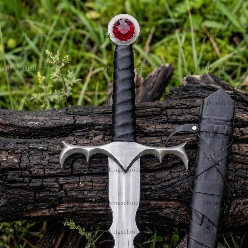 May include: A steel sword featuring a black leather-wrapped grip, a silver crossguard with pointed ends, and a circular pommel set with a faceted red gemstone. A matching black leather scabbard with crisscross lacing is positioned beside the sword.