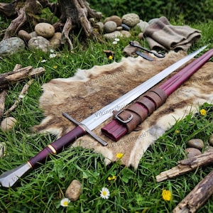Puede incluir: Una espada de estilo medieval con empuñadura burdeos y hoja plateada, sobre una piel. La espada tiene una vaina de cuero con hebilla. La escena está ambientada al aire libre sobre hierba verde.