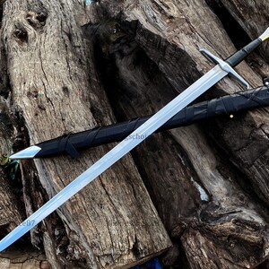 May include: A long, silver sword with a black handle and crossguard, resting on a wooden surface. The sword is sheathed in a black scabbard. The blade has a sharp point and a polished finish. The handle has a gold accent.