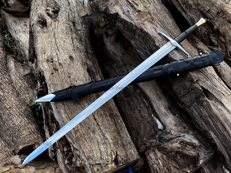 May include: A long, silver sword with a black handle and crossguard, resting on a wooden surface. The sword is sheathed in a black scabbard. The blade has a sharp point and a polished finish. The handle has a gold accent.