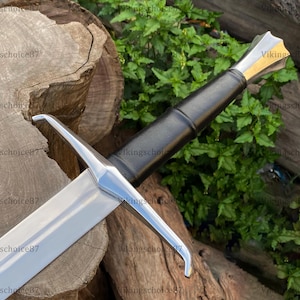 May include: A medieval sword with a silver blade and a black leather-wrapped handle. The hilt features a gold accent. The sword is displayed on a wooden surface with green foliage in the background.