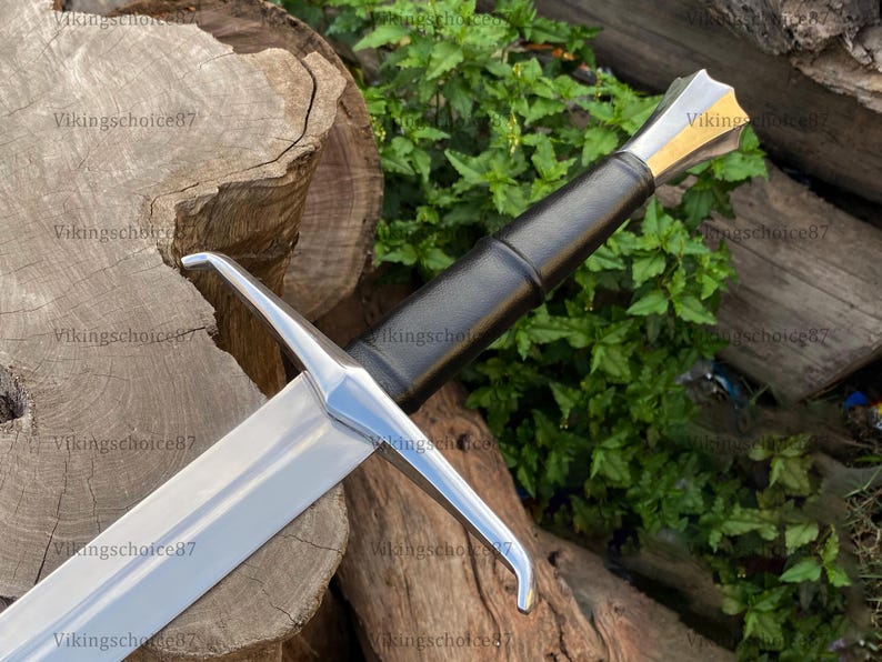 May include: A medieval sword with a silver blade and a black leather-wrapped handle. The hilt features a gold accent. The sword is displayed on a wooden surface with green foliage in the background.