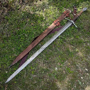 May include: A long, ornate sword with a patterned blade and a brown leather-wrapped handle. The sword rests on green grass next to its wooden sheath, which is also brown and has leather straps.