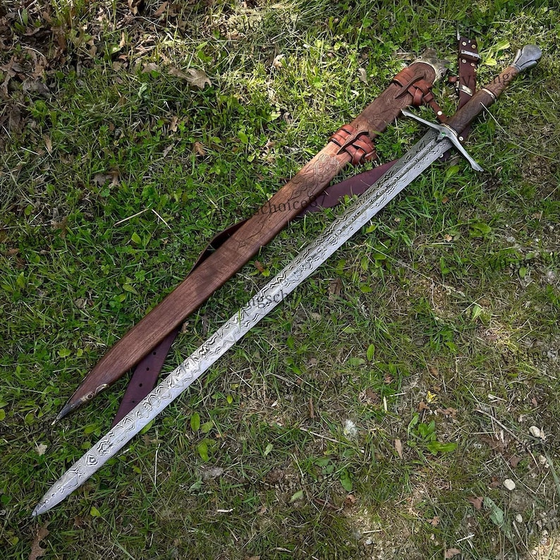 May include: A long, ornate sword with a patterned blade and a brown leather-wrapped handle. The sword rests on green grass next to its wooden sheath, which is also brown and has leather straps.