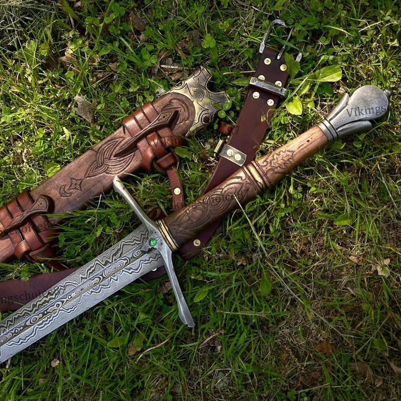 May include: A detailed image of a Viking sword with a patterned blade and wooden handle. The sword rests on green grass next to a brown leather sheath and belt. The handle is engraved with intricate designs, and the blade has a unique pattern.