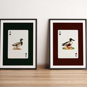 Peut inclure: Deux impressions d'art encadrées représentant des cartes à jouer. Chaque impression affiche un As avec une illustration détaillée d'un canard. Une carte a un fond vert foncé et l'autre un fond bordeaux. Les cadres sont noirs.