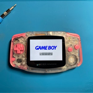 Puede incluir: Una consola de juegos portátil Game Boy Advance transparente rosa con un logotipo "GAME BOY" azul en la pantalla. La pantalla también muestra "BRT CLR S/P F/P" y "14 2 2 0H".