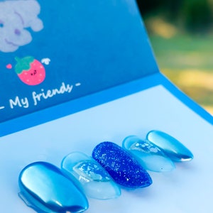May include: A set of six blue press-on nails in various shades and finishes, including a glittery accent nail. The nails are displayed on a white surface with a blue box featuring a cartoon strawberry and text that reads "My friends -."