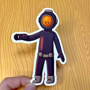 May include: A sticker featuring a cartoon diver in a dark purple diving suit with a round orange face and a smiling face. The diver is wearing a helmet and boots, and has a black arm extended. The sticker is on a wood grain background.