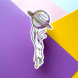 Puede incluir: Una pegatina holográfica de una mano sosteniendo un planeta con anillos. La mano gotea una sustancia blanca.