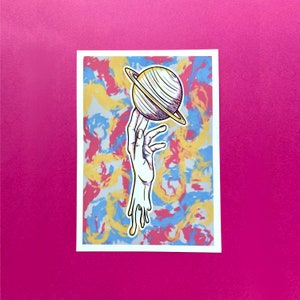 Puede incluir: Una mano con pintura goteando sostiene un planeta amarillo y blanco con un anillo a su alrededor. El fondo es un diseño abstracto colorido con tonos de rojo, amarillo, azul y gris.