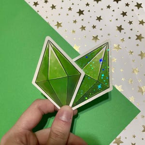 以下が含まれることがあります： 白い縁取りのある、緑色のダイヤモンド型のステッカーが2つ。1つのステッカーには、青と緑のキラキラ光るホログラフィック効果があります。ステッカーは、金色の星が散りばめられた緑と白の背景にあります。