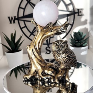 May include: A gold-colored decorative owl figurine perched on a tree branch, holding a light pink crystal ball. The figurine is set against a white background with a black compass rose and small potted plants.