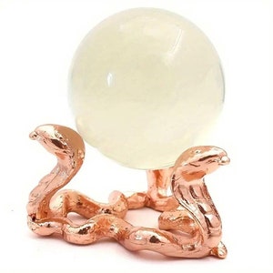 May include: A clear, glowing orb rests on a rose gold-colored stand. The stand is crafted in the shape of two snakes, their bodies intertwined. The orb appears to be made of glass or a similar translucent material.