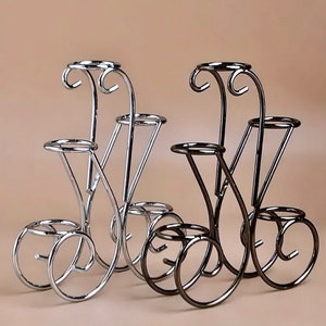 May include: Two decorative metal plant stands. One is silver, the other is dark bronze. Each stand has three circular platforms of varying sizes, connected by curved and straight metal supports. The top of each stand has a decorative scroll.