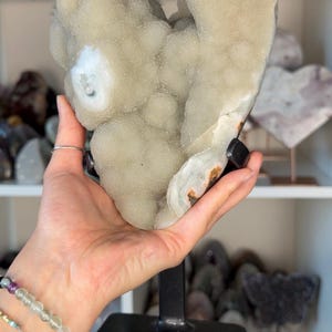 May include: A large, light-colored geode crystal formation with a textured surface, mounted on a black metal stand. The crystal has a rounded shape with a few darker spots. The background shows other crystals on a shelf.