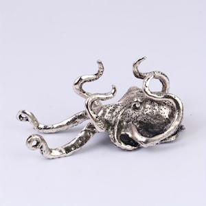 Könnte beinhalten: Eine detaillierte, silberfarbene Oktopus-Figur mit strukturierter Haut und gekräuselten Tentakeln. Die Skulptur ist vor einem schlichten weißen Hintergrund platziert, der ihr kompliziertes Design und das maritime Thema hervorhebt. Das Objekt ist dekorativ.