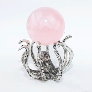 May include: A light pink rose quartz sphere displayed on a silver-colored octopus-shaped stand. The stand has detailed tentacles that cradle the sphere. The background is plain white.