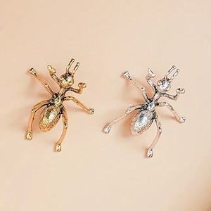 May include: Two metal ant-shaped jewelry pieces. One is gold-toned, the other silver-toned. Both have detailed bodies, six legs, and antennae. The ants are positioned on a light peach background, showcasing their intricate design.