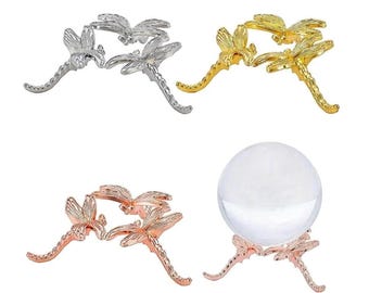 May include: Four decorative dragonfly-shaped stands in silver, gold, and rose gold. One stand holds a clear crystal ball. The stands are made of metal and have detailed dragonfly wings and bodies. The stands are designed to hold spheres.