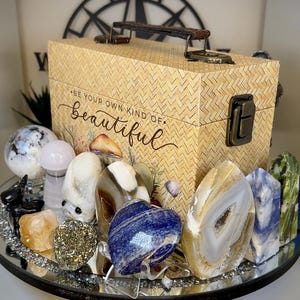 May include: A collection of polished gemstones and crystals, including a blue heart-shaped stone, agate, and quartz, arranged on a mirrored tray. A woven box with the text "BE YOUR OWN KIND OF beautiful" is in the background.