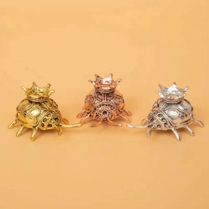 Op de afbeelding: Drie decoratieve keverbeeldjes in goud, brons en zilver. Elke kever heeft een kroon op zijn kop en gedetailleerde patronen op zijn schild. De beeldjes worden gepresenteerd op een effen, lichtoranje achtergrond.