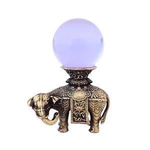 May include: A decorative bronze-colored elephant figurine with intricate detailing, supporting a glowing, lavender-colored orb. The elephant has a detailed saddle and trunk raised. The orb is smooth and spherical, creating a mystical aesthetic.