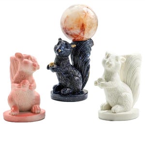 May include: Three squirrel figurines in different colors. One is pink, another is black with a sphere, and the third is white. The black squirrel holds a sphere. The figurines are on a white background.