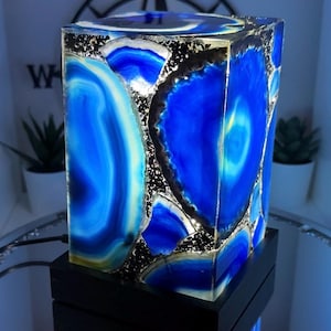 May include: A rectangular lamp with striking blue agate slices, featuring white and black detailing. The lamp is set on a black base, reflecting on a mirrored surface. The agate design creates a vibrant, illuminated effect.