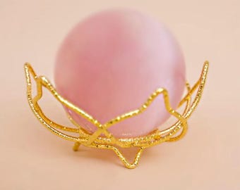 May include: A pink sphere sits in a gold-coloured, decorative metal holder. The holder has a stylised, openwork design with a textured surface. The background is a soft, neutral colour.