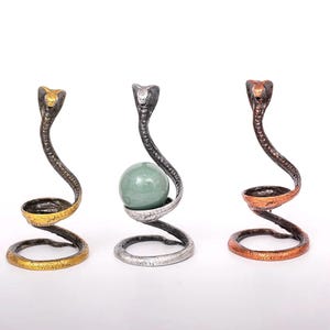 May include: Three metal cobra sculptures in gold, silver, and copper tones. The cobra's body forms a spiral base and supports a small bowl or a green sphere. The sculptures have a detailed, textured finish.