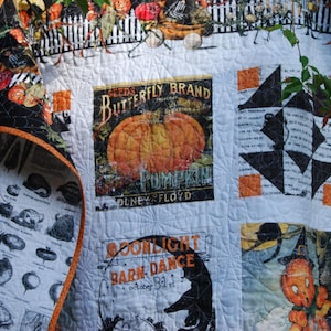 May include: A white quilt featuring various Halloween-themed panels, including a large orange pumpkin with the text Seeds Butterfly Brand Pumpkin, a black cat silhouette with the text Moonlight Barn Dance, and a black and white botanical print.