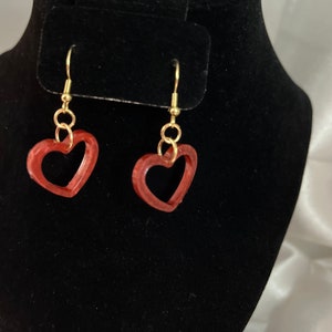 Könnte beinhalten: Ein Paar goldene herzförmige Ohrringe mit einem roten, durchsichtigen, offenen Herzdesign.