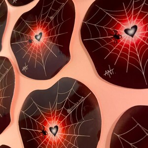 May include: A set of six black and red spiderweb stickers with a white heart and spider in the center. Each sticker has the text "ANT" and "@amaya.sart" printed on it.