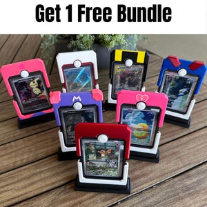 May include: A collection of seven trading card display stands shaped like miniature ball devices, each holding a different collectible card. The stands feature various color schemes including pink, white, black, yellow, blue, purple, and red. Text reads Buy 3 Get 1 Free Bundle.