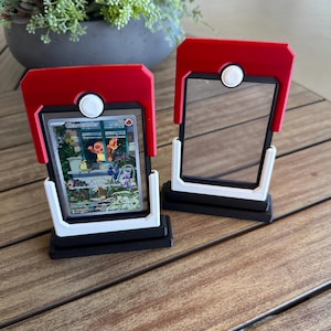 Könnte beinhalten: Zwei rechteckige Sammelkarten-Präsentationsständer im Design eines Pokeballs. Jeder Ständer verfügt über einen roten oberen Teil mit einem weißen runden Knopf, einen schwarzen Rahmen und einen weißen Sockel. Einer enthält eine Charmander-Sammelkarte.