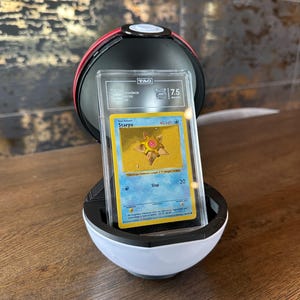May include: A graded Staryu trading card in a clear plastic slab, featuring a yellow star-shaped character on a blue background, displayed inside an open red, white, and black spherical container that resembles a classic ball from the game series.