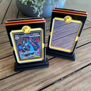 Könnte beinhalten: Zwei schwarze Aufsteller mit goldenen und roten Akzenten, jeweils mit einem rechteckigen Rahmen. Einer enthält eine Mega Charizard X-Sammelkarte mit japanischem Text, während der andere leer ist und eine reflektierende Fläche zeigt.