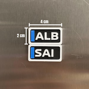 K&ouml;nnte beinhalten: Zwei rechteckige Magnete mit wei&szlig;em Rand, schwarzer Mitte und blauen vertikalen Streifen auf der linken Seite. Der obere Magnet zeigt den wei&szlig;en Text ALB, der untere den wei&szlig;en Text SAI. Jeder Magnet misst 4 cm mal 2 cm.
